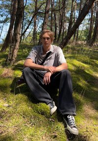 Giovane seduto su muschio verde in una foresta, indossando una camicia a quadri e pantaloni neri, con sneakers nere e una borsa a tracolla nera.