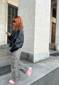 Zwart leren jack, broek met luipaardprint, roze sneakers. Staand naast een stenen zuil, met een smartphone in de hand, en golvend rood haar.