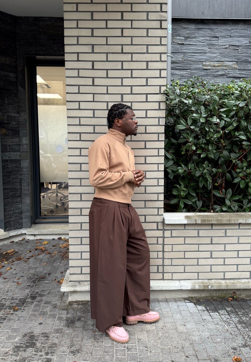 Sweat-shirt beige à fermeture éclair, pantalon large marron, et chaussures épaisses roses. Debout contre un mur en briques, avec de la verdure en arrière-plan.