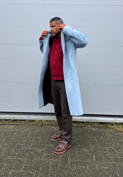 Abrigo largo azul claro, suéter de punto burdeos, pantalones de cuadros marrones y zapatos marrones con cordones. Texturas suaves y corte relajado.