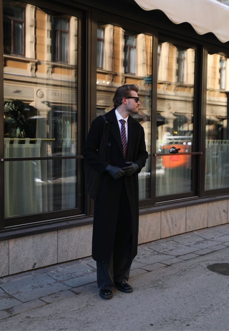 Homme en manteau noir, gants et lunettes de soleil, debout sur le trottoir de la ville devant des fenêtres de bâtiment réfléchissantes, regardant vers la droite.