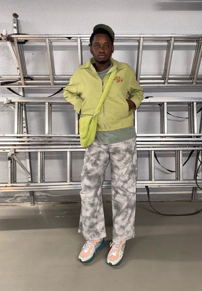 Chaqueta verde clara con detalles bordados, camiseta gris, pantalones de pierna ancha de tie-dye y zapatillas multicolores. Pequeño bolso cruzado verde de peluche.