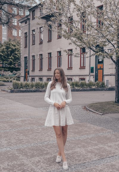 Suéter blanco de manga larga y un vestido ligero de corte acampanado. De pie sobre un pavimento de adoquines con un edificio gris claro y vegetación de fondo.