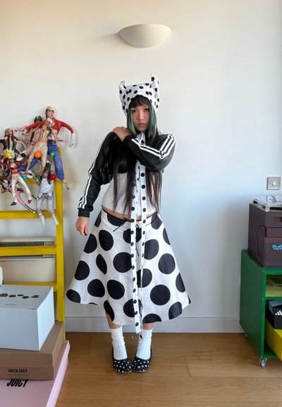 Persona con falda de lunares blancos y negros, sombrero a juego, chaqueta negra, de pie dentro de una habitación con estanterías y figuras de muñecas.