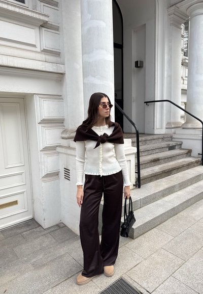 Mujer con blusa blanca abotonada y pantalones anchos oscuros, de pie cerca de las escaleras de un edificio con columnas blancas, sostiene un bolso negro y lleva gafas de sol.