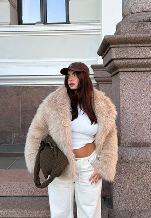 Mujer con abrigo de piel sintética beige, top corto blanco, pantalones blancos, gorra marrón y sosteniendo un bolso tejido verde oliva, de pie junto a un edificio de piedra.