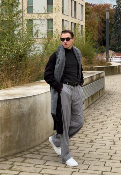 Abrigo negro, bufanda gris, camiseta negra ajustada, pantalones grises holgados y zapatillas blancas. Entorno urbano con vegetación y un fondo de edificios modernos.