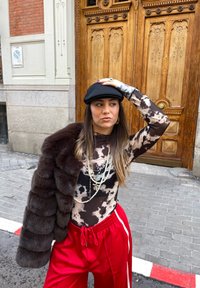 Cappello piatto nero, top trasparente a maniche lunghe con motivi marroni e bianchi, giacca in pelliccia sintetica, collane di perle sovrapposte, pantaloni rossi con strisce bianche.