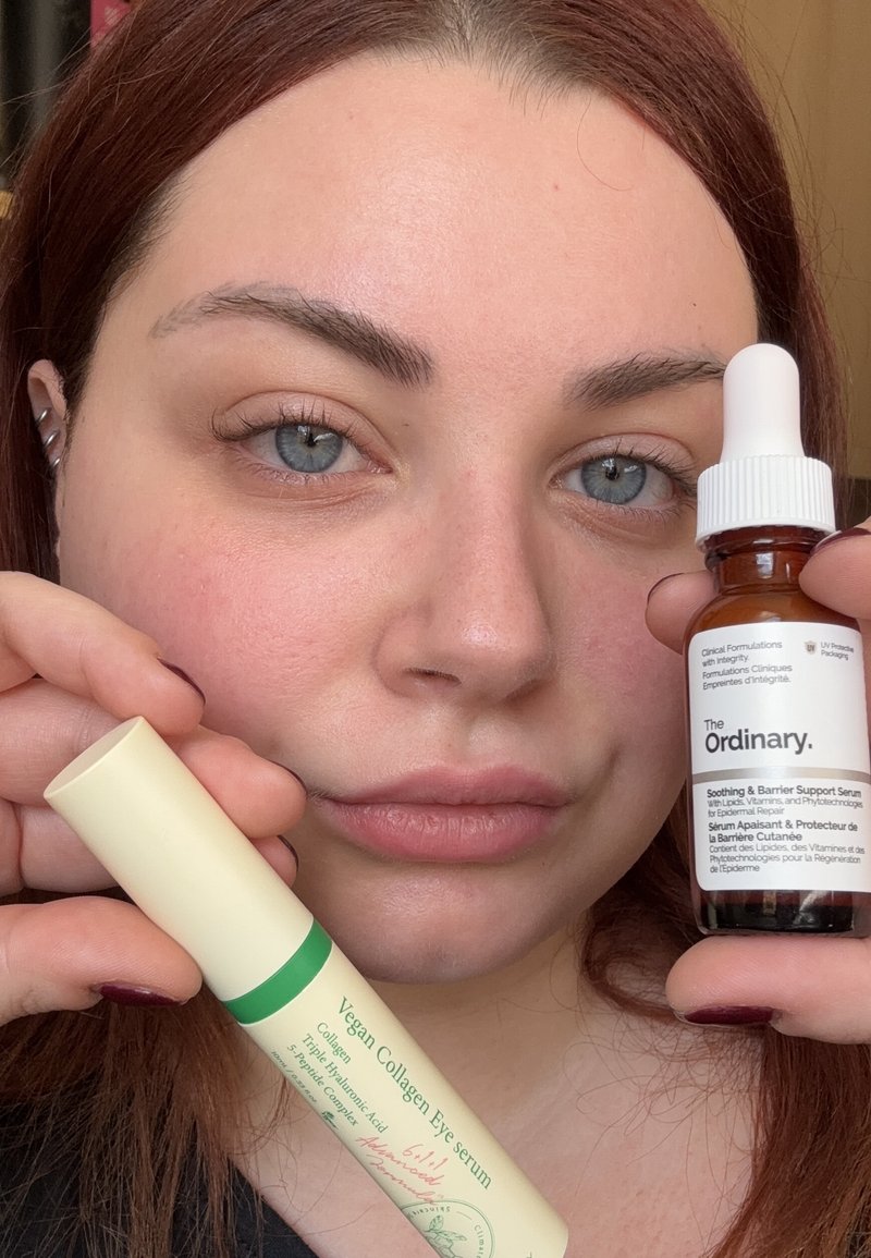 Glass dropperflaske med The Ordinary serum med hvid hætte og brunt væske, ved siden af et cylindrisk beige rør med grønne accenter til vegansk øjenserum.
