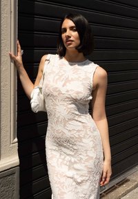 Biała koronkowa sukienka z kwiatowym wzorem, dopasowany fason i bezrękawowy krój. Połączona z małą białą torebką. Czarny tło.