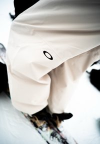 Pantaloni da neve beige chiaro realizzati in materiale resistente all'acqua, con un logo circolare nero sulla gamba e una vestibilità ampia per la massima libertà di movimento.