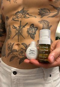 Due prodotti per la cura della pelle tenuti contro un addome tatuato: una bottiglia bianca con contagocce etichettata "The Ordinary" e una bottiglia spray verde etichettata "ELEMIS Superfood".