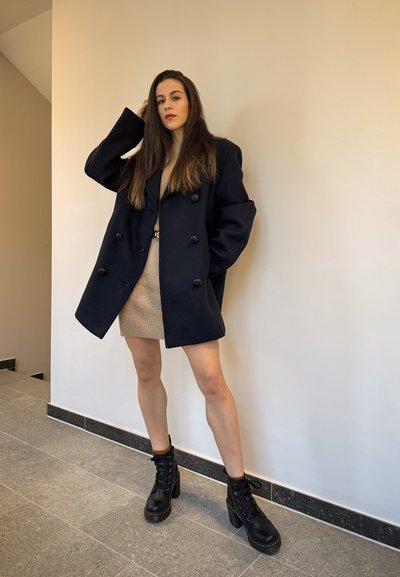 Abrigo navy de doble botonadura, vestido de punto beige y botas de tacón negro con cordones. El entorno es un interior con azulejos y paredes en tonos neutros.