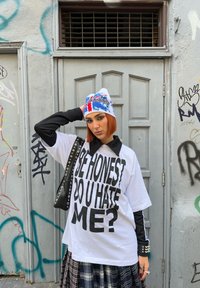 Maglietta bianca oversize con testo nero, maniche lunghe nere e gonna a quadretti. Presenta un berretto blu e bianco con design grafico.