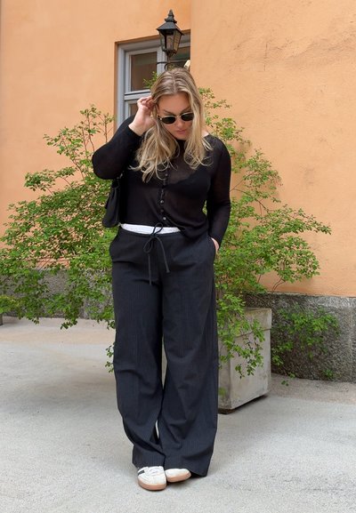 Top negro transparente de manga larga con botones, combinado con pantalones de pierna ancha a rayas y zapatillas blancas. Fondo de follaje verde y pared naranja.