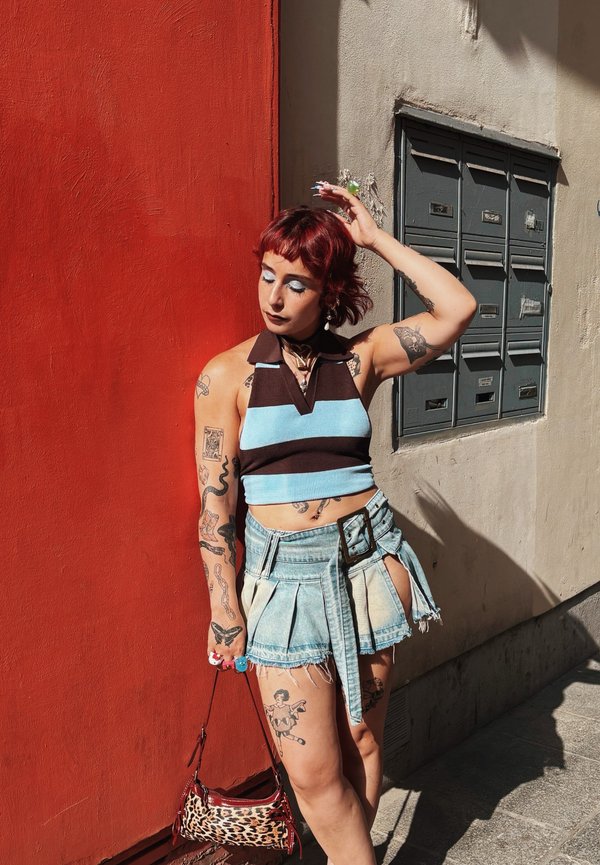 Donna con capelli rossi che indossa un crop top a righe e una gonna di jeans, in piedi contro un muro rosso alla luce del sole, che tiene una borsa con stampa leopardata.