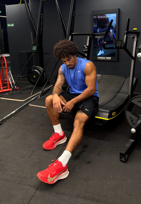 Hombre con camiseta sin mangas azul y zapatos rojos sentado en una cinta de correr del gimnasio, descansando con la cabeza baja y las manos en las rodillas.