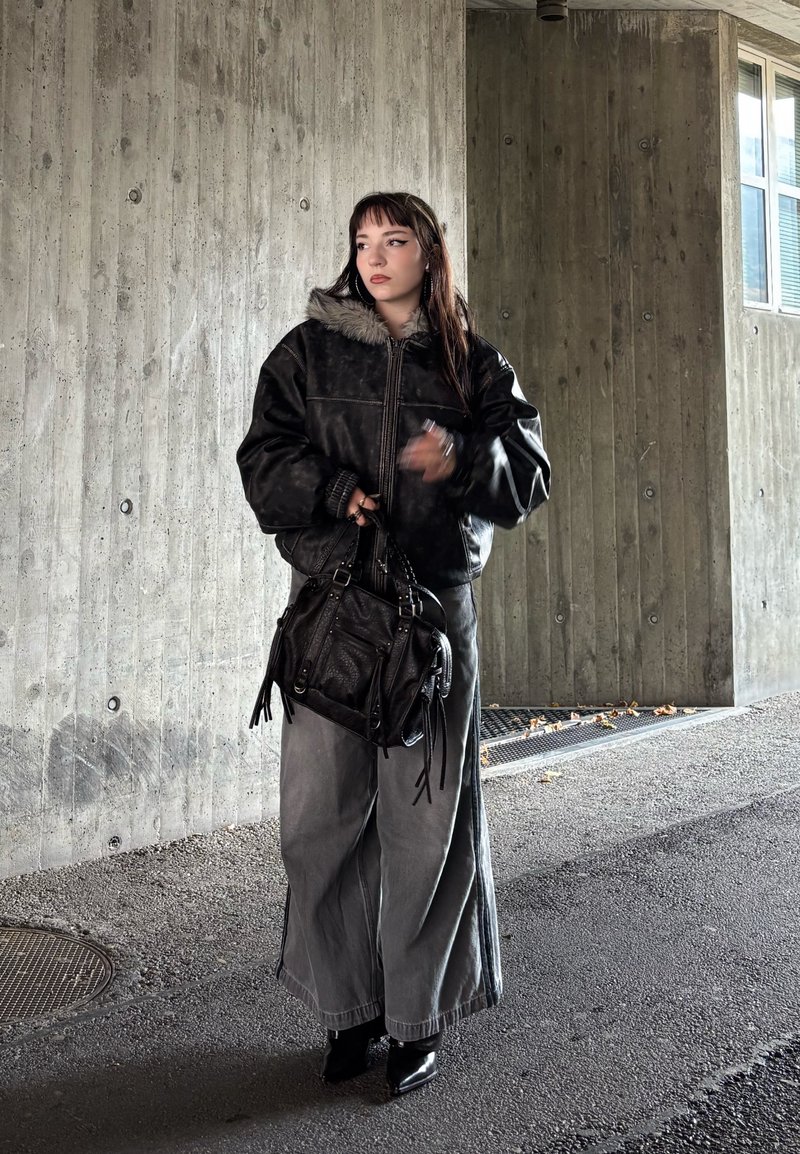 Frau in schwarzer Jacke mit Pelzkragen und weiten grauen Hosen, die eine schwarze Handtasche hält und vor einer Betonwand in der Nähe eines Fensters steht.