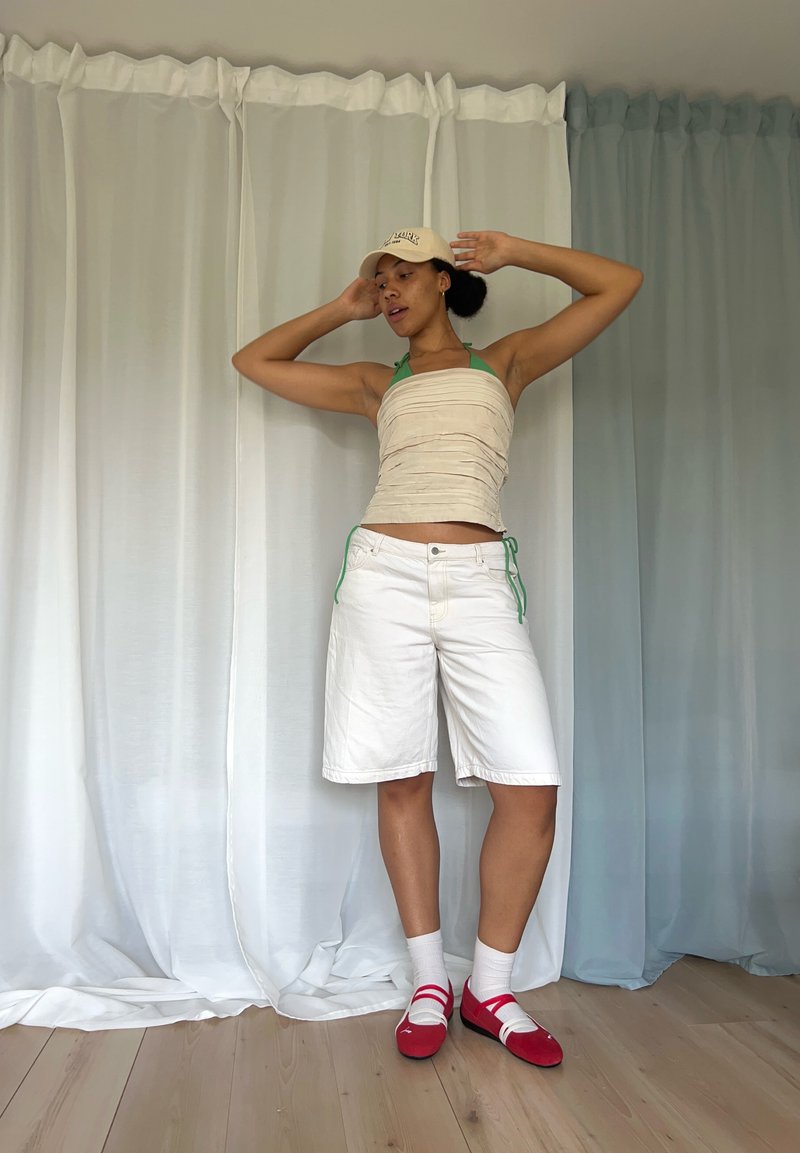 Jovem posando com as mãos perto da cabeça, vestindo uma blusa bege, calções brancos, sapatos vermelhos com meias brancas e um boné bege contra um fundo de cortinas.