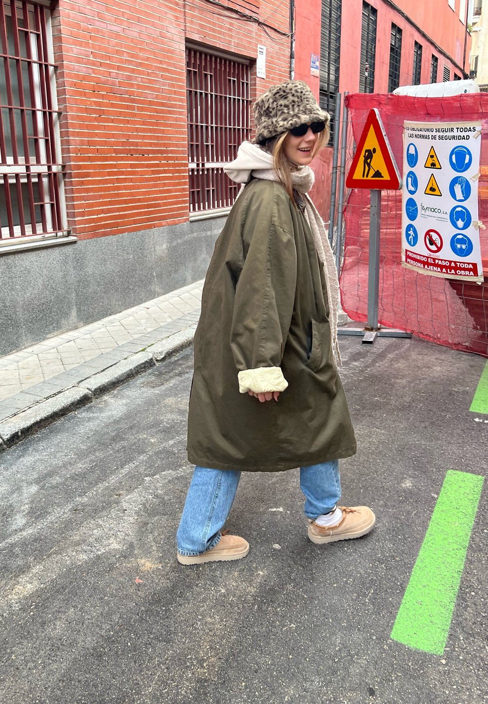 Casaco oversized verde-oliva com punhos acolchoados claros, moletom cinza por baixo, jeans azuis e ténis bege. Chapéu de padrão leopardo e óculos de sol.
