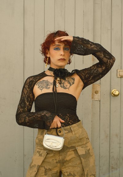 Persona con cabello rizado rojo que lleva una blusa de encaje negra, pantalones de camuflaje, sosteniendo un pequeño bolso plateado, de pie frente a una puerta gris.