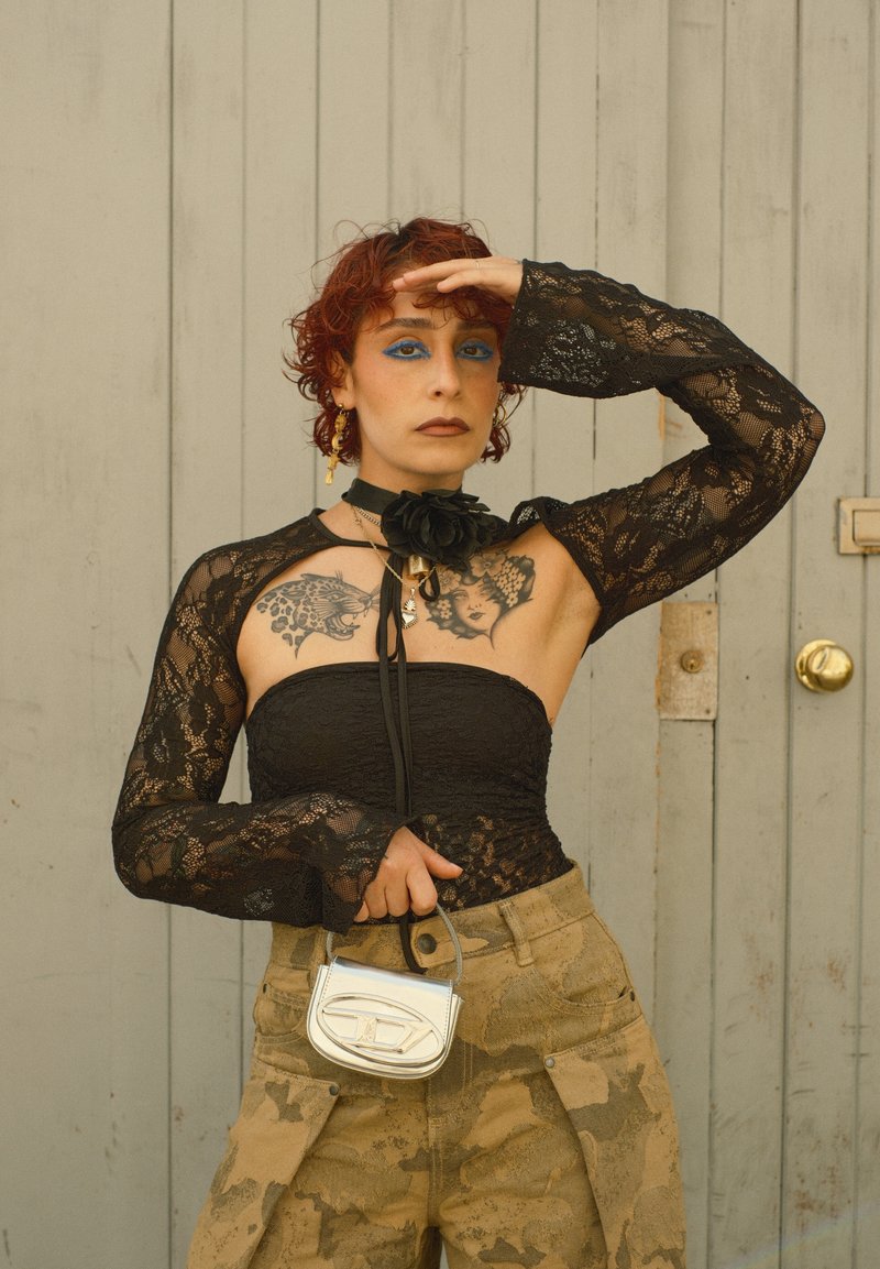 Personne aux cheveux roux bouclés portant un haut en dentelle noire, un pantalon camouflage, tenant un petit sac à main argenté, debout devant une porte grise.