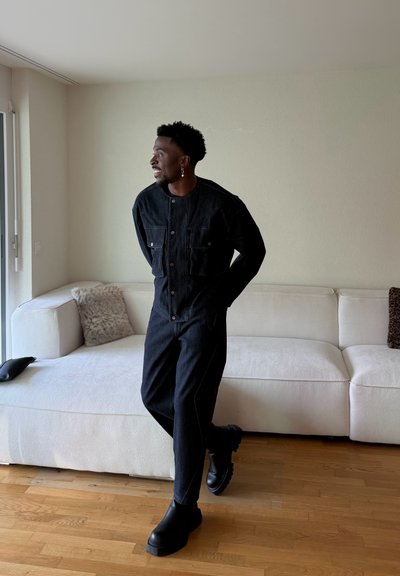 Hombre con atuendo de mezclilla negro y botas, de pie en la sala junto a un sofá blanco, mirando por la ventana con las manos detrás de la espalda.