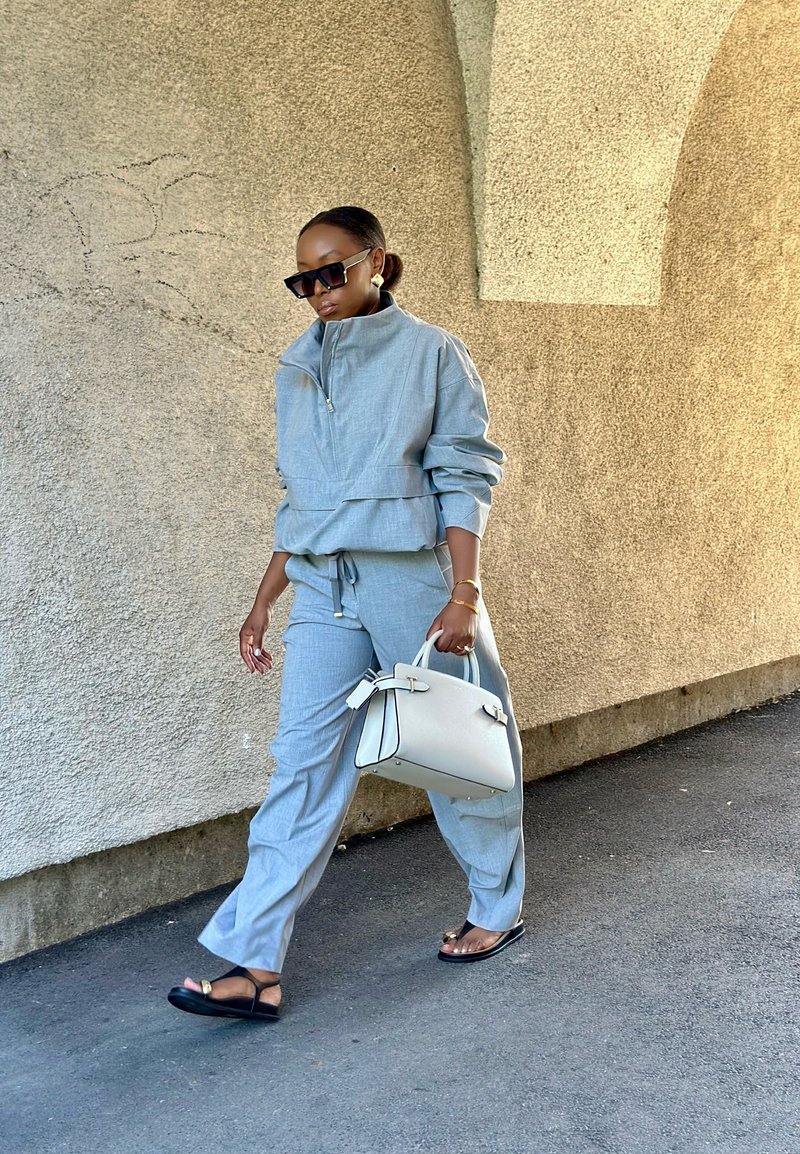 Completo athleisure grigio chiaro con giacca a zip e pantaloni con coulisse; abbinato a una borsa bianca e sandali neri. Texture liscia.