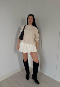 Bež pulover z pletenjem, bele kratke puffy hlače, črne kolenske škornje in črna torbica na ramenu. Gladke teksture in kontrastne barve.