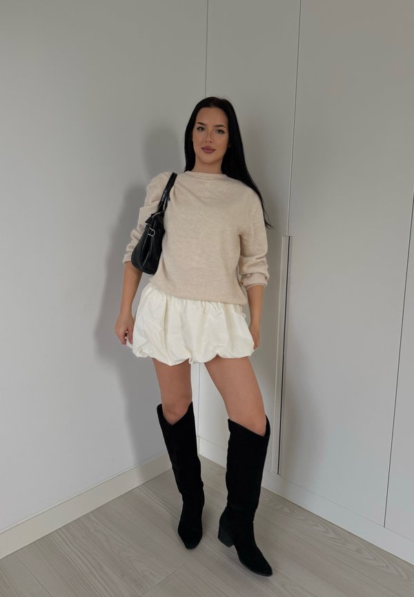 Pull en tricot beige, shorts blancs bouffants, bottes noires montantes et un sac à épaule noir. Textures lisses et couleurs contrastées.