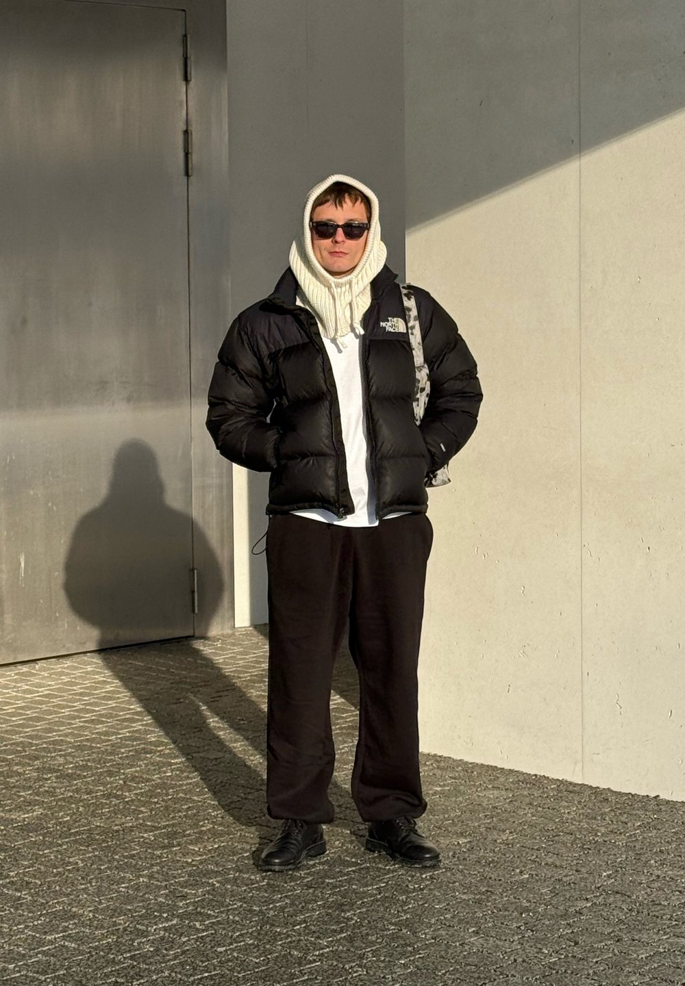 Personne portant une doudoune noire, un pantalon noir, un bonnet en tricot blanc à capuche, des lunettes de soleil et des chaussures noires, debout sur un pavé texturé près d'une porte métallique et d'un mur.