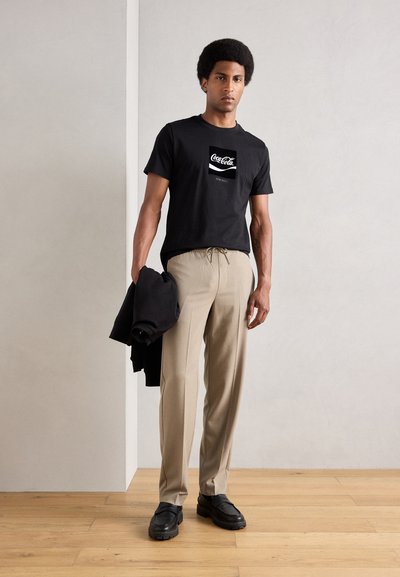 Camiseta de algodón negra con gráfico de Coca-Cola, pantalones plisados beige y zapatos negros sin cordones. Modelo sosteniendo una chaqueta negra. Suelo de madera.