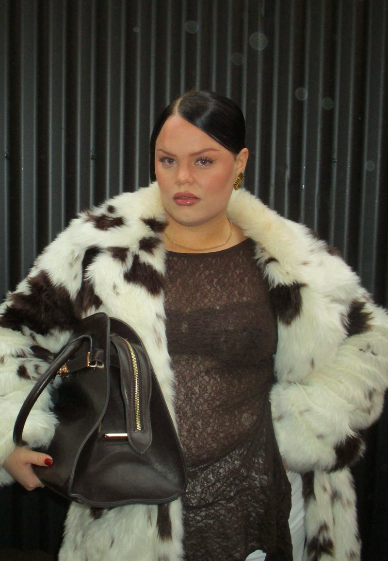 Manteau en fausse fourrure blanche et marron, top marron transparent, boucles d'oreilles créoles dorées, tenant un sac à main marron foncé avec des accents de zip doré. Fond texturé.