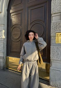 Krøllet kvinde i grå træningsdragt står ved en stor trædør, holder solbriller og justerer sit hår, med olivengrøn taske over skulderen.