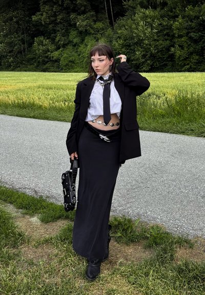 Blazer negro sobre una camisa blanca con una corbata negra, combinado con una falda larga negra. Accesorios plateados, un bolso negro con tachuelas y botines.