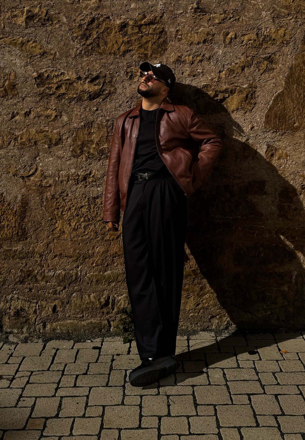Chaqueta de cuero marrón sobre una camisa negra, combinada con pantalones negros sueltos y zapatillas negras. De pie contra una pared de piedra texturizada.