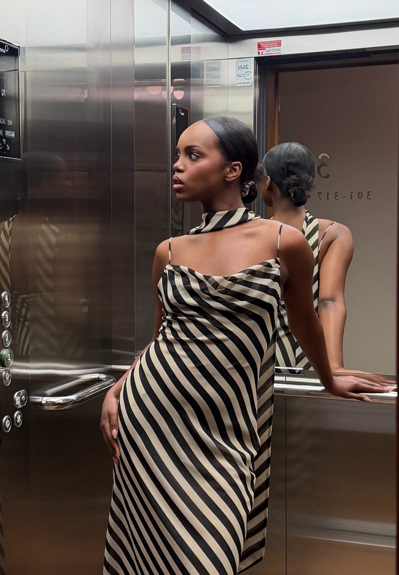 Mujer con vestido a rayas negras y beige apoyada en la barandilla dentro de un ascensor de metal reflectante, mirando hacia su izquierda.