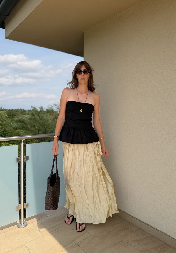 Frau mit schwarzem trägerlosem Oberteil, langem beigen Rock, Sonnenbrille und Flip-Flops steht auf dem Balkon und hält eine geflochtene Tasche.