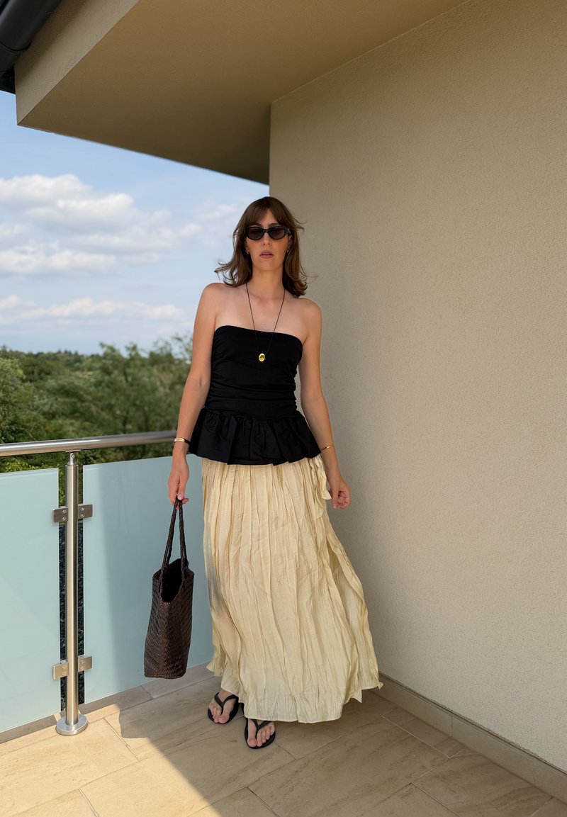 Frau mit schwarzem trägerlosem Oberteil, langem beigen Rock, Sonnenbrille und Flip-Flops steht auf dem Balkon und hält eine geflochtene Tasche.