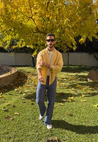 Άνδρας με γυαλιά ηλίου και κίτρινο πουκάμισο που σηκώνει τα μανίκια, περπατώντας σε χλόη κάτω από ένα δέντρο με φωτεινά κίτρινα φύλλα σε έναν ηλιόλουστο κήπο.