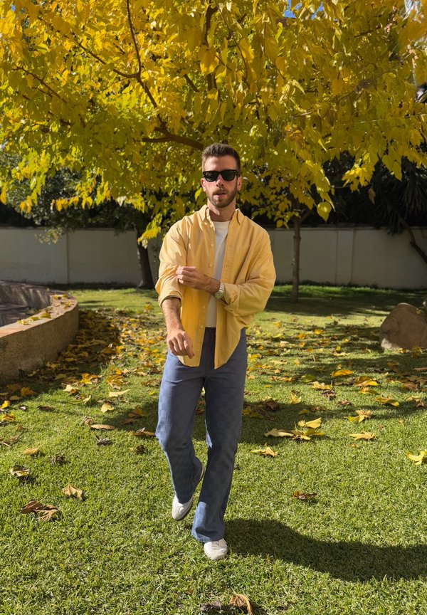 Vyras su akiniais nuo saulės ir geltona palaidine susukantis rankoves, vaikščioja ant žolės po medžiu su ryškiai geltonais lapais saulėtoje sode.