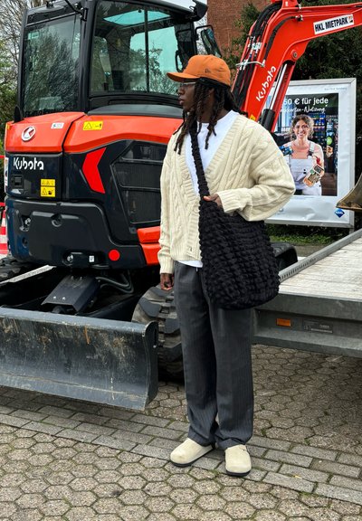 Cárdigan de punto en color beige claro, camisa blanca, bolsa tote negra con textura, pantalones a rayas grises y zapatos tipo slip-on beige junto a una excavadora naranja.