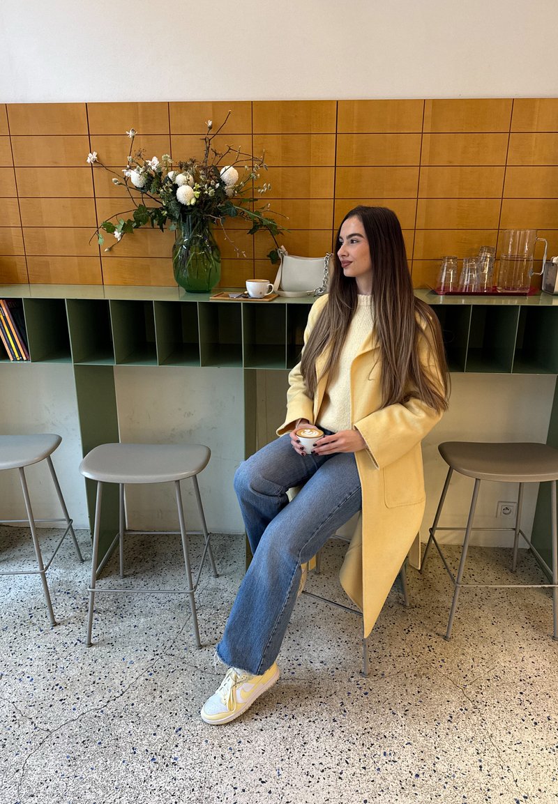 Gul uldfrakke over beige sweater, blå jeans med vidde og lyse sneakers. Sidder ved en grøn disk med en blomsterdekoration og en kop.