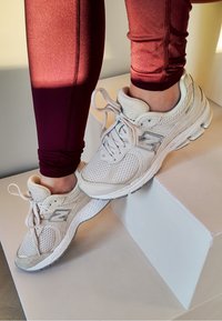 Light grey New Balance trainers made from mesh and suede, featuring a thick white sole and round laces, placed on a white step.