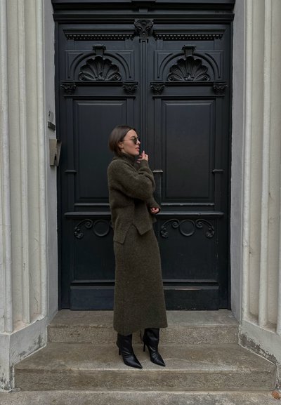 Un conjunto de lanas texturizadas en verde oscuro que consta de un suéter de cuello alto y una falda larga, complementado con botas negras de punta afilada, cerca de una puerta negra ornamentada.
