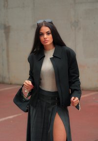 Črn zip-up jopič preko bež rebraste majice, v kombinaciji s sivo karo krilo z visokim razporkom in zaponkami. Prisoten je tudi majhen črn ročaj.