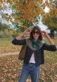 Cardigan grigio con motivo geometrico bianco, sciarpa verde, t-shirt bianca, jeans blu e occhiali da sole marroni. Borsa verde oliva a spalla, foglie autunnali.