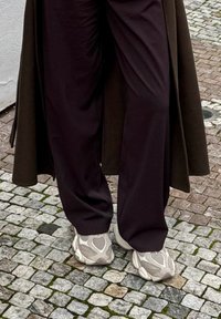 Personne portant un pantalon ample sombre et des baskets gris clair surdimensionnées, debout sur un pavement en pavés.