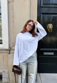 Hvid langærmet skjorte med løs pasform, lyseblå brede jeans og en lille mørkebrun crossover-taske. Tekstureret stenvæg og løve dørhåndtag.