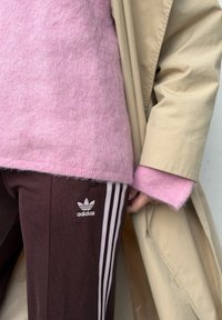 Rosa fluffy genser lagd under en beige jakke, sammen med brune bukser med tre hvite striper og et Adidas-logo.